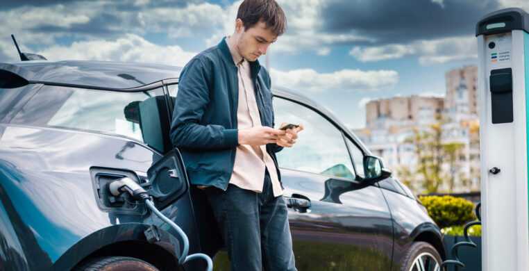 Man charging his electric car at charge station and using smartphone