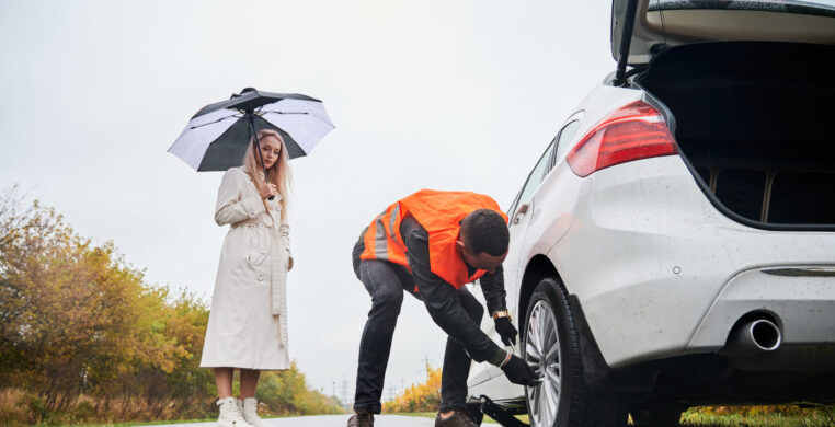 Male auto mechanic unscrewing lug nuts on car wheel while elegant woman holding umbrella. Young man in vest repairing woman automobile on the road. Concept of emergency road service.